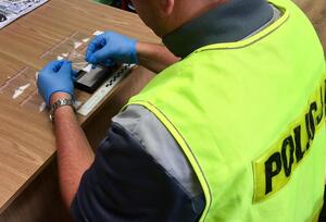 Policjant siedzący przy biurku, który ma założone rękawiczki jednorazowe i w ręku trzyma woreczki z narkotykami.