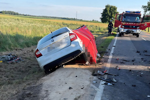 Biały samochód osobowy po wypadku stoi przechylony w przydrożnym rowie. Obok pojazdu rozstawiono czerwony parawan zasłaniający miejsce działań ratowniczych. Na jezdni leżą odłamki karoserii, a w tle widoczny jest wóz strażacki i strażak.
